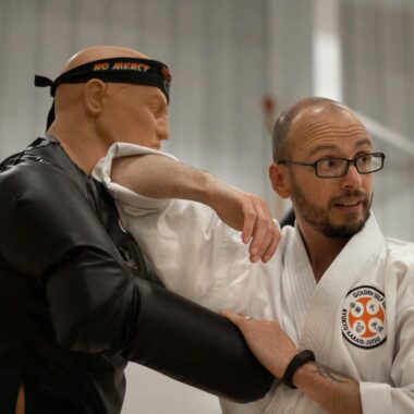 An instructor teaches self-defense using a martial arts dummy indoors.